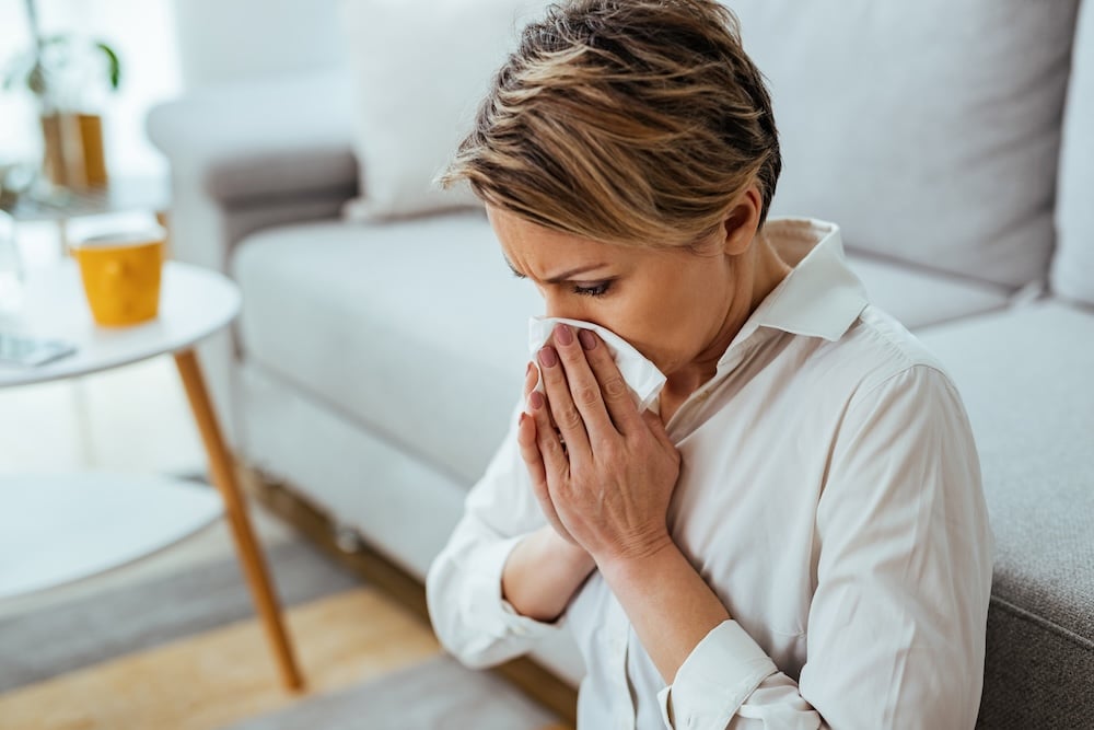 Businesswoman having a flue and blowing nose in a tissue at home. Griep.