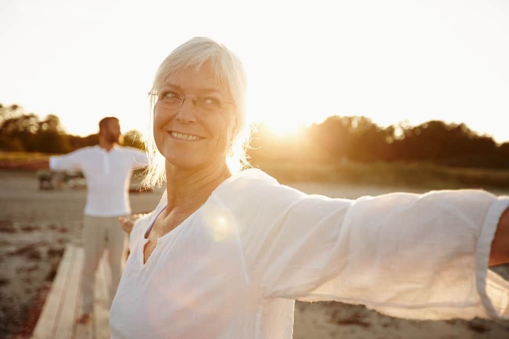 Every day is a yoga day. Shot of a mature woman doing yoga on the beach with her husband in the background.