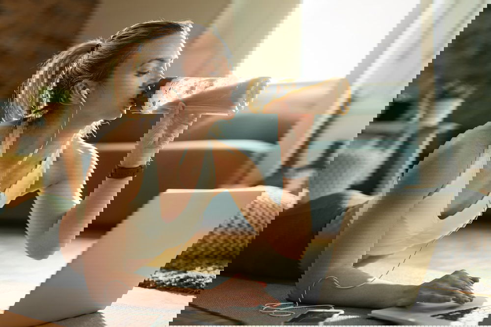 Eiwitten - Young athletic woman drinking protein shake while using laptop on the floor.